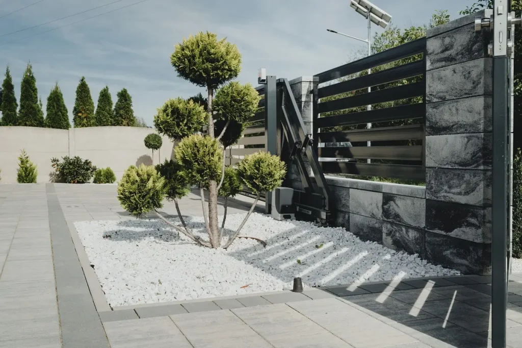 Modernne ogrodzenie z automatyczną bramą i ozdobnym drzewkiem.